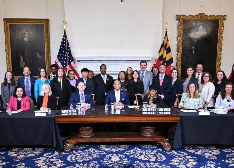 Maryland Governor, Wes Moore sits in the middle of a large table. He is surrounded by witnesses to the signing.