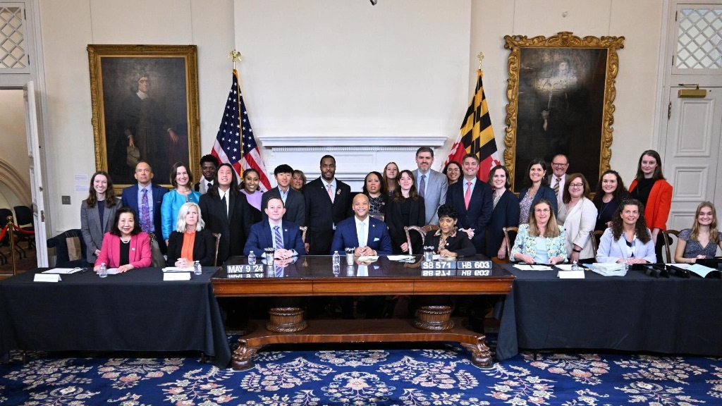 Maryland Governor, Wes Moore sits in the middle of a large table. He is surrounded by witnesses to the signing.