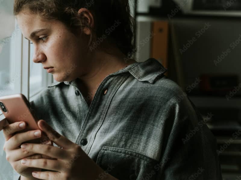A one-quarter headshot of a teen girl in a plaid shirt holding a pink-cased smartphone. She is standing next to a closed window looking solemn. You can see her reflection in the window. The feel of the image is sad.