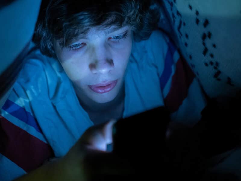 A young boy is using a smartphone while lying in bed under a blanket at night. The light of the phone screen is bouncing off his hand and face.