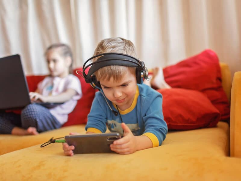 A young boy is lying down on a yellow sofa. He is lying on his front with his space position towards the camera. He is wearing headphones and looking intently at a phone. A young girl is in the background, on the sofa, on a laptop, but out of focus.