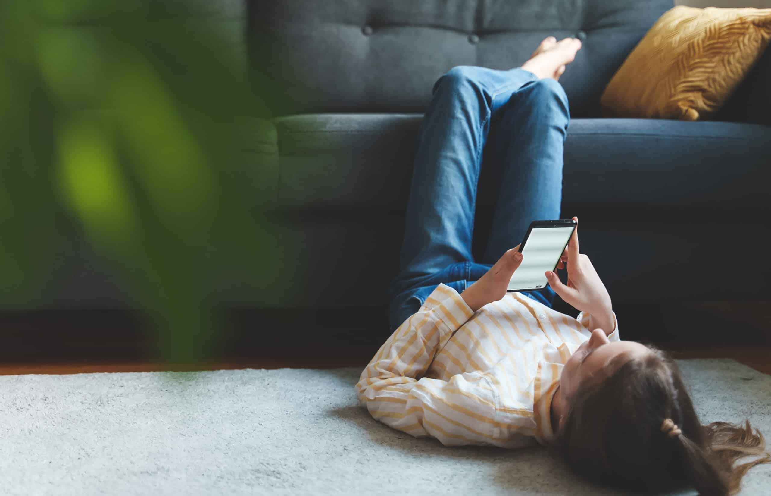 Teen girl with smartphone relaxing at home. She is lying on the floor, with legs resting on the sofa. There is an out-of-focus plant in the forground, to the left of the image.