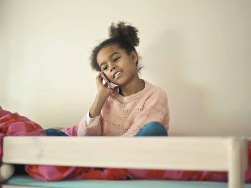 A black girl, sat on her bed on top of her duvet. She is holding a smartphone in right hand and holding it to her ear. Her head is tilted to the left and she is smiling.