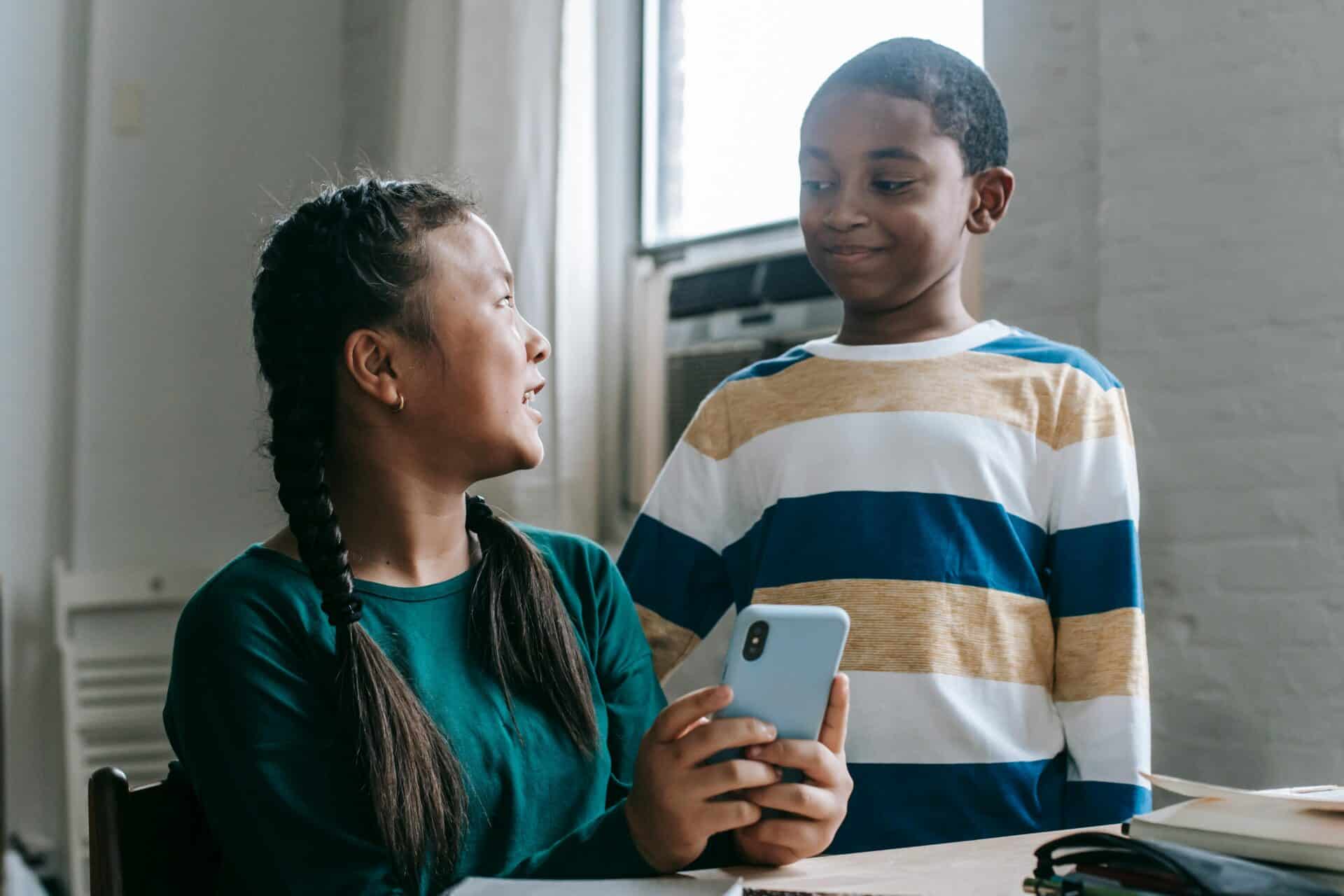 A young girl with black hair pleated in braids and wearing a teal long-sleeve shirt is showing something on her phone to her friend, a boy seemingly younger than her wearing a striped long-sleeve shirt. The boy is standing while she is sitting down.