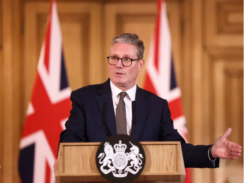 UK Prime Minister Keir Starmer delivers a speech from behind a wooden podium adorned with the royal coat of arms. The setting features two large British flags in the background and a formal interior with wooden paneling.