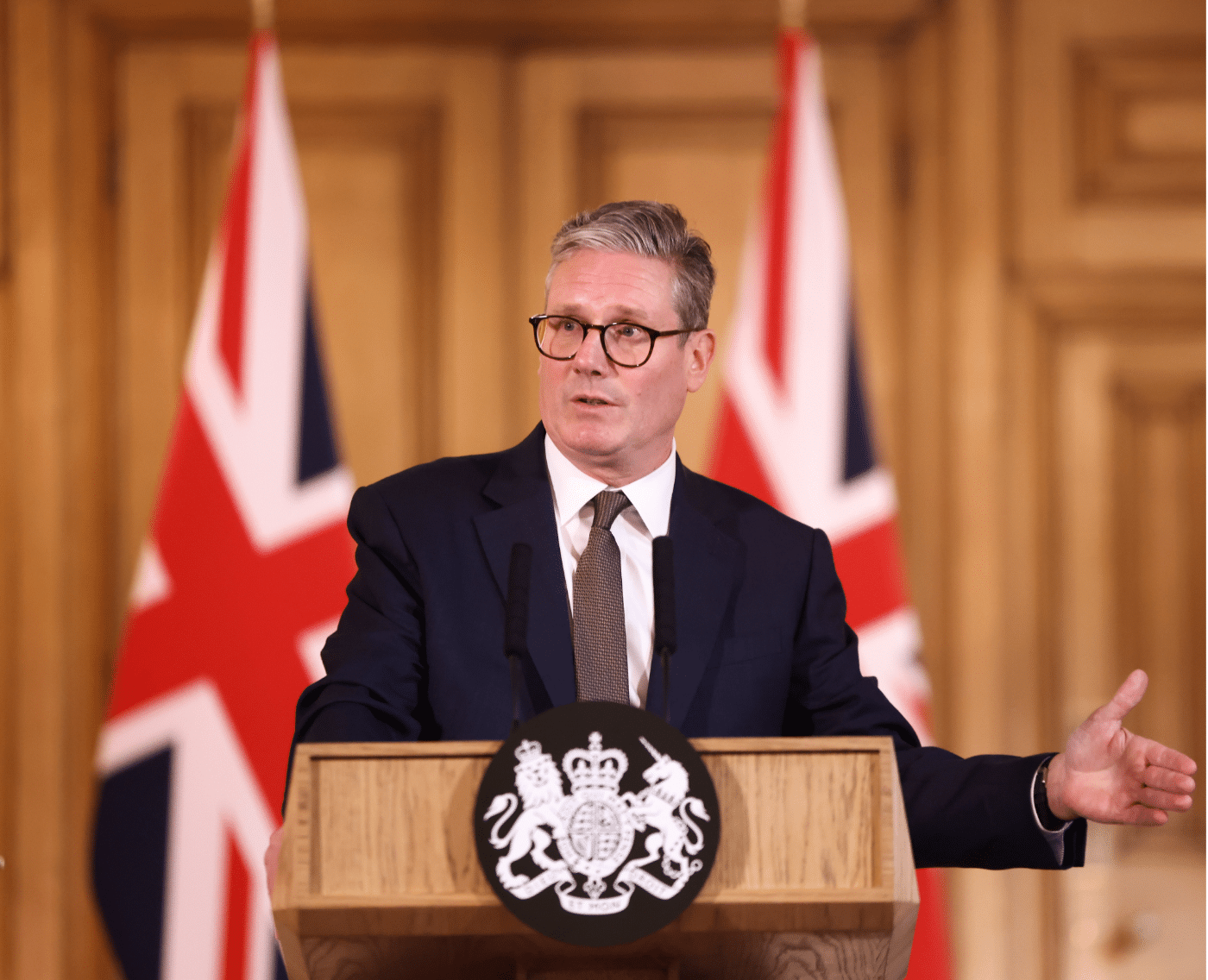 UK Prime Minister Keir Starmer delivers a speech from behind a wooden podium adorned with the royal coat of arms. The setting features two large British flags in the background and a formal interior with wooden paneling.