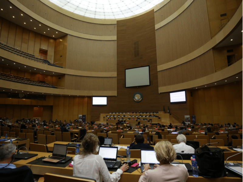 A full view of the conference room at the Global Summit to Tackle Online Sexual Exploitation. The large room features tiered seating with participants working on laptops. At the front is a podium and a large screen. The high ceiling and skylight brighten the space, adding to the formal yet collaborative environment of the summit.