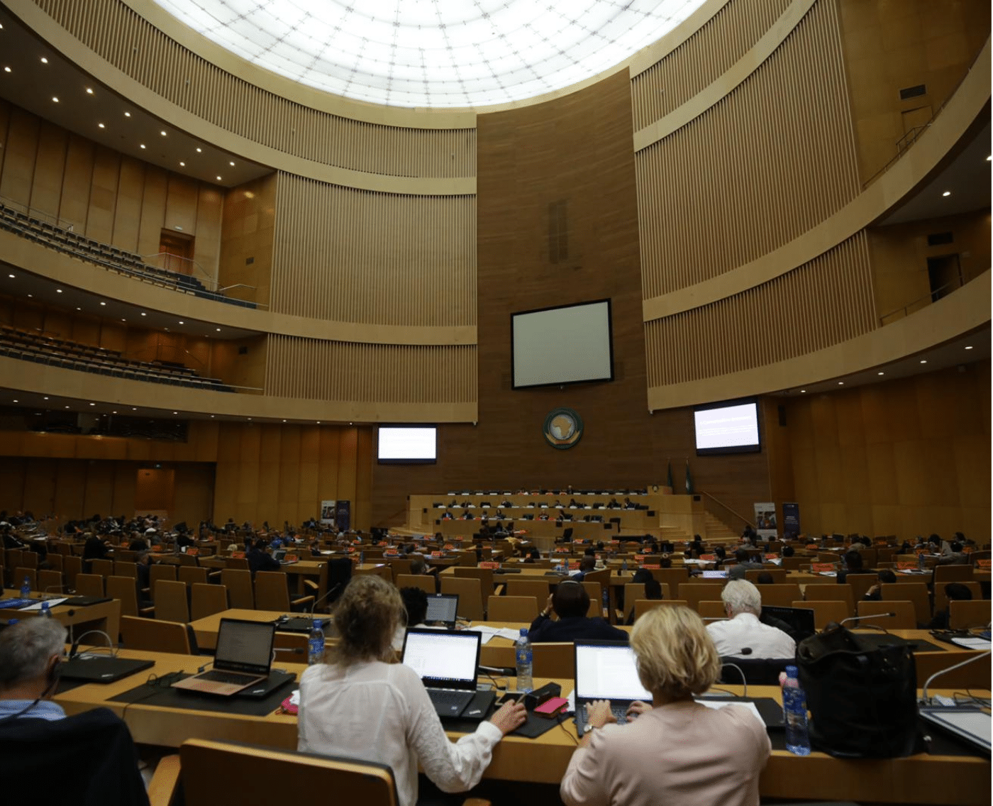 A full view of the conference room at the Global Summit to Tackle Online Sexual Exploitation. The large room features tiered seating with participants working on laptops. At the front is a podium and a large screen. The high ceiling and skylight brighten the space, adding to the formal yet collaborative environment of the summit.