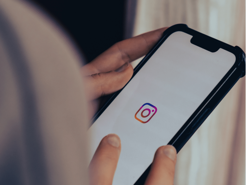 A wider shot showing a person holding a smartphone with the Instagram logo displayed on the screen. The person’s hands are visible as they hold the phone, and the screen is in focus, while the background is blurred. This is the full version of the close-up shown above.