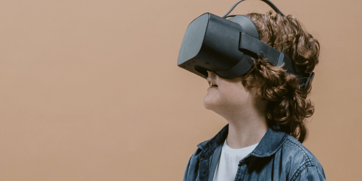 A young child with curly hair is wearing a virtual reality headset and smiling. They are dressed in a denim jacket and a white shirt, standing against a plain beige background.
