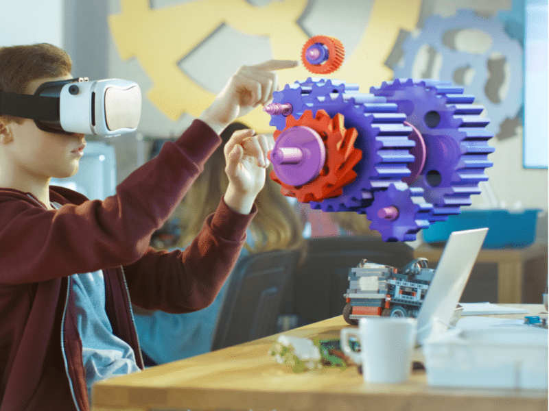 A young boy wearing a virtual reality headset is engaged in an interactive learning experience. He is sitting at a desk in a classroom, and with his hands, he appears to be manipulating a holographic 3D model of colourful gears floating in the air in front of him. The gears are large, vividly coloured in purple, red, and blue, and seem to be part of a mechanical assembly. The background shows other students working.