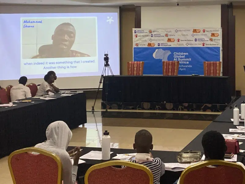A conference room. On two projector screens is a video testimony from a young Ghanaian person. Sitting on tables in the foreground, facing away from the camera and looking towards the screen is three African children.