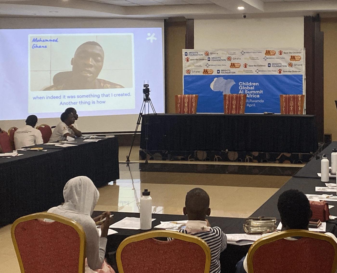 A conference room. On two projector screens is a video testimony from a young Ghanaian person. Sitting on tables in the foreground, facing away from the camera and looking towards the screen is three African children.