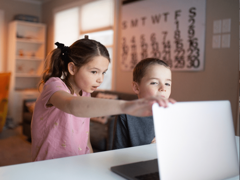 Two children are sitting at a table, focused on a laptop. The girl points at the screen while the boy looks on attentively. The background shows a cosy room with a yellow chair, a bookshelf, and a large calendar hanging on the wall.