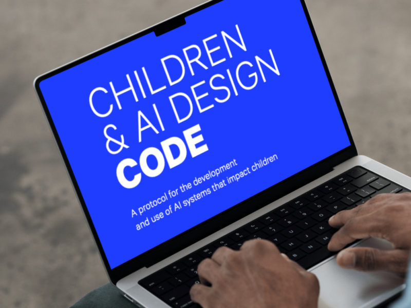 Top black hands type on a laptop. The laptop is angled towards the screen. On the screen, it reads Children & AI Design Code in white on a bright blue background.