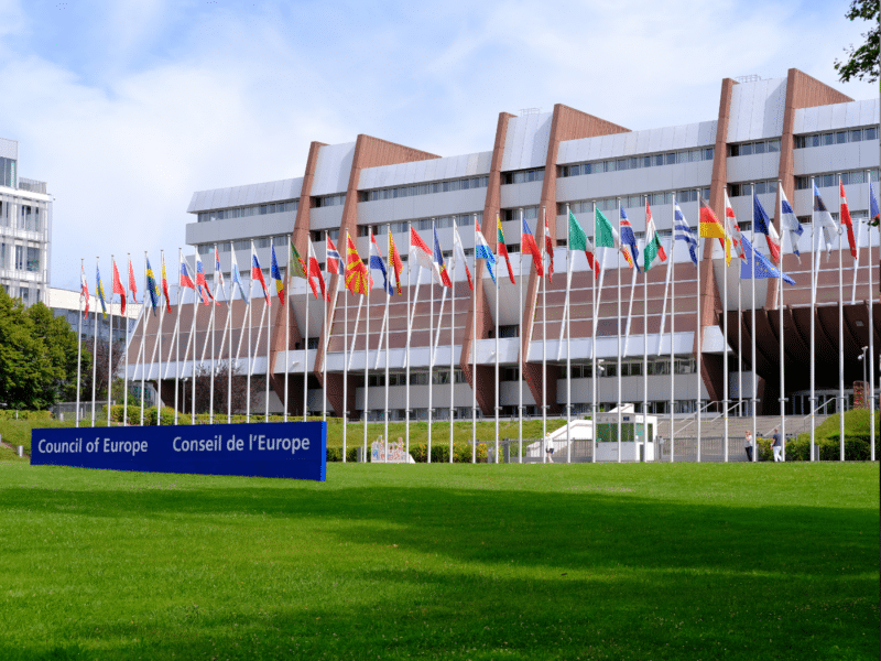 A wide shot of the Council of Europe building in Strasbourg, France. The building is a large, modern structure with a flat roof and a lawn in front of it. Many flags are flying in front of the building, representing the different member states of the Council of Europe. The sky is blue with some white clouds.