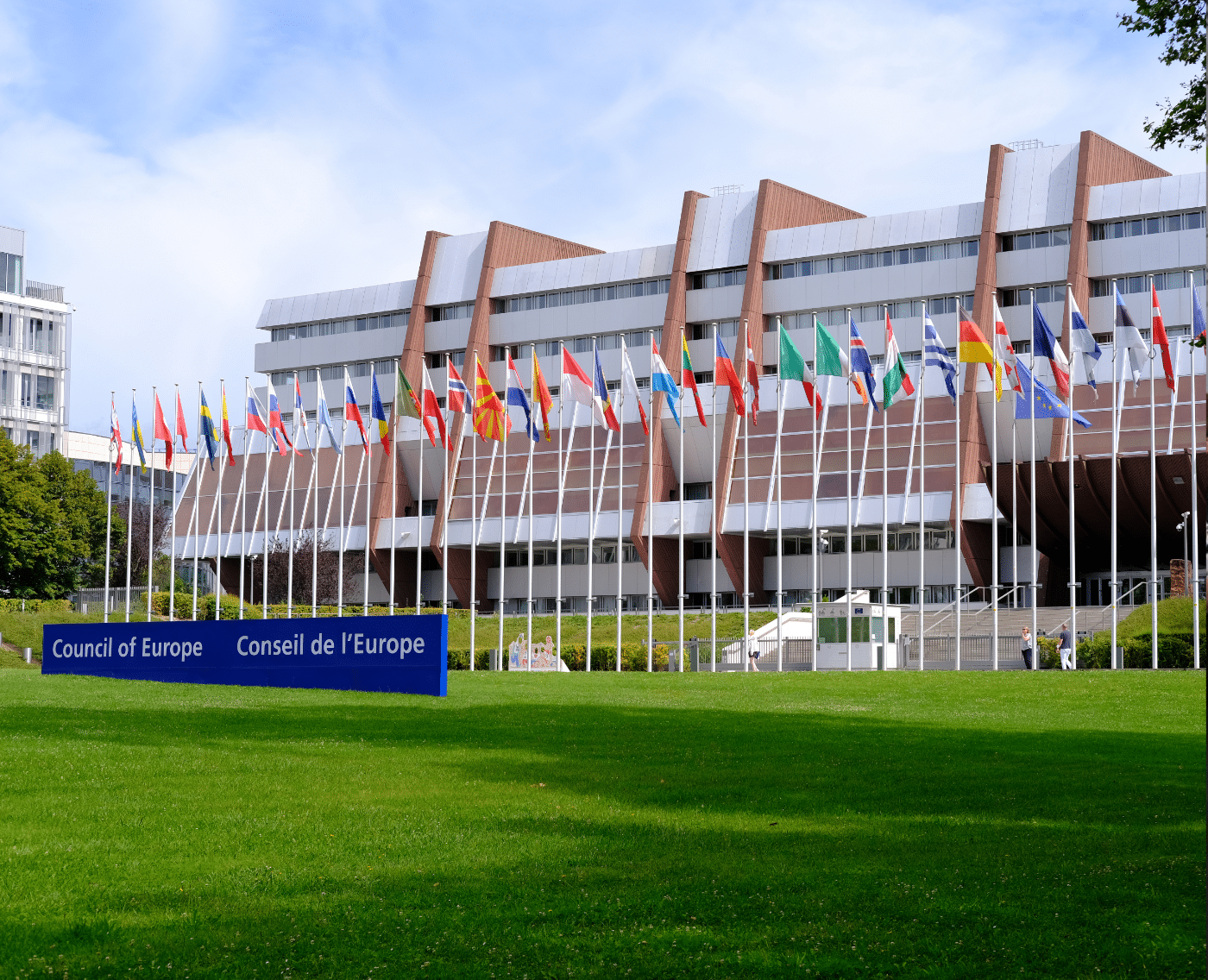 A wide shot of the Council of Europe building in Strasbourg, France. The building is a large, modern structure with a flat roof and a lawn in front of it. Many flags are flying in front of the building, representing the different member states of the Council of Europe. The sky is blue with some white clouds.