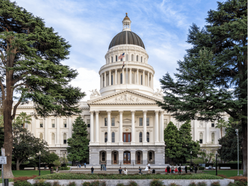 A photograph of the California State Capitol building in Sacramento. The neoclassical architecture features a large central dome, flanked by columns and a pediment at the entrance. The dome is topped with a smaller structure and an American flag flying at the peak. The building is surrounded by lush greenery, including tall trees that frame the scene on both sides.