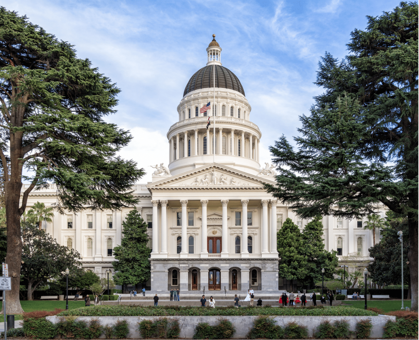 A photograph of the California State Capitol building in Sacramento. The neoclassical architecture features a large central dome, flanked by columns and a pediment at the entrance. The dome is topped with a smaller structure and an American flag flying at the peak. The building is surrounded by lush greenery, including tall trees that frame the scene on both sides.