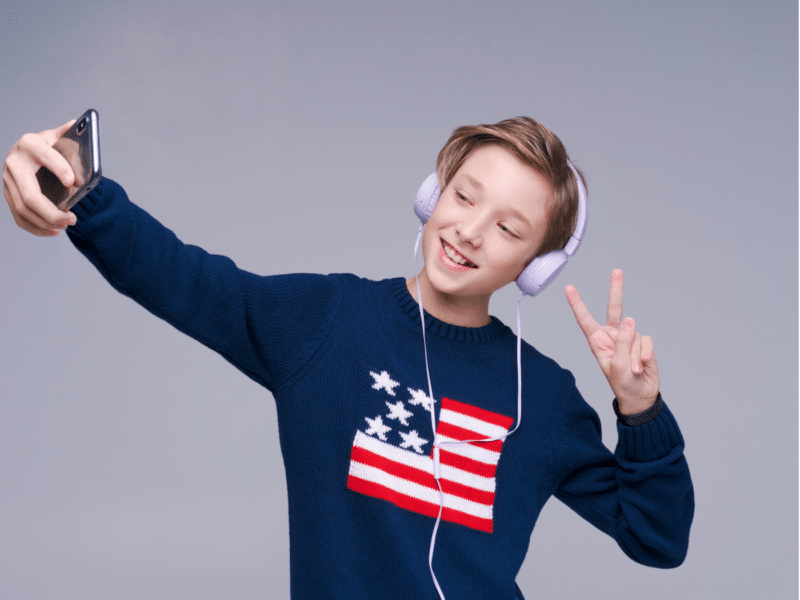 A young boy taking a selfie with a smartphone while wearing light purple headphones. He is smiling and holding up a peace sign with his other hand. The boy is wearing a dark blue sweater with an American flag design on the front.