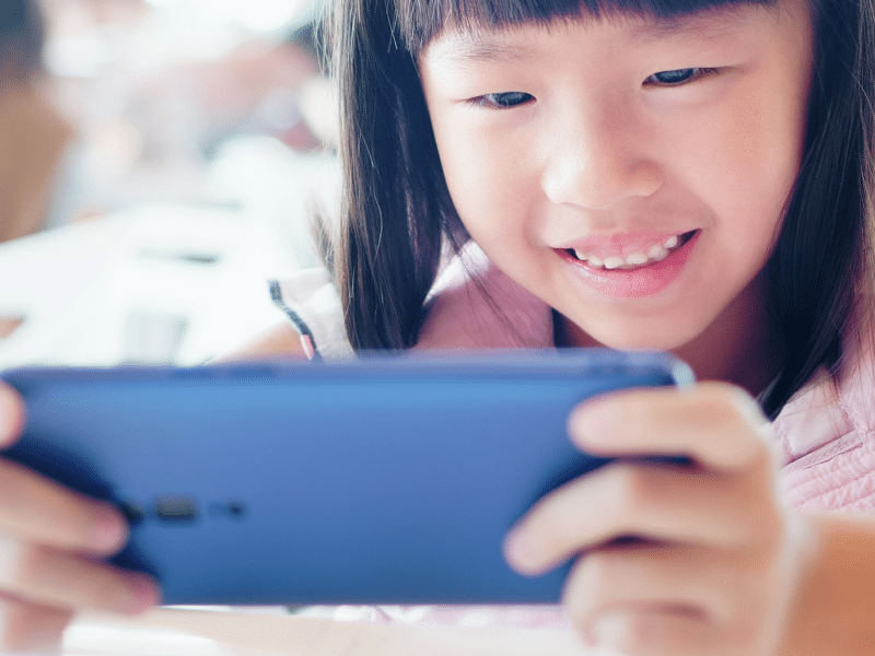 A young girl with straight black hair is smiling as she looks at a smartphone she is holding with both hands. The focus is on her face and the phone, suggesting she is enjoying a game or video. The background is blurred.