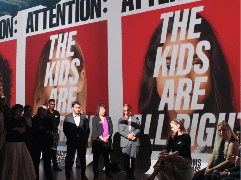 A group of people standing and sitting in front of large posters with bold text that reads "ATTENTION: THE KIDS ARE ALL RIGHT" on a red background. The posters feature partially obscured images of children's faces, with the text overlaid on them. The group is at a conference in Toronto titled “Attention: The Kids Are All Right”, with some standing in a row while others sit, facing toward the camera. The setting suggests a formal or professional gathering, with a spotlight on a few attendees and the posters in the background dominating the space.