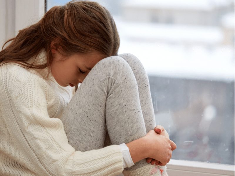 A full view of the young girl sitting in the same position by the window. The girl is hugging her knees, with her head resting on them.