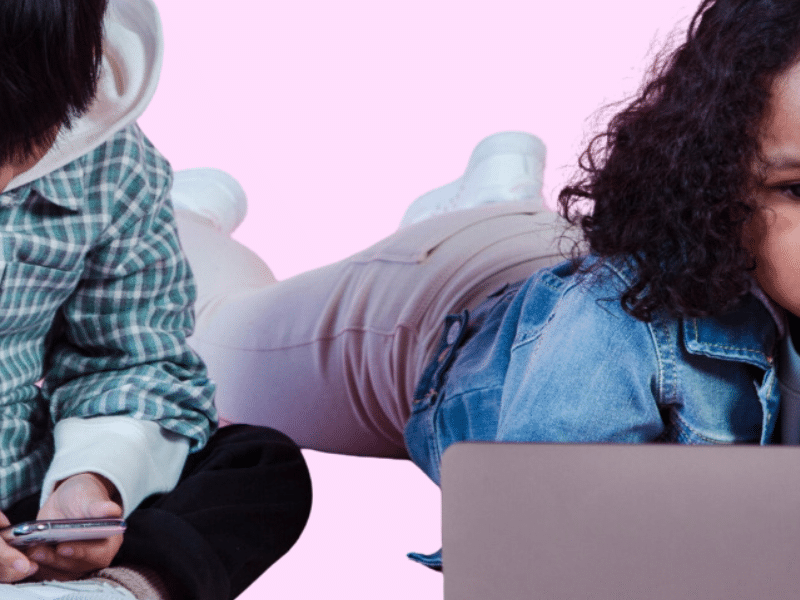 Two children are laying down on the floor staring at their respective devices.