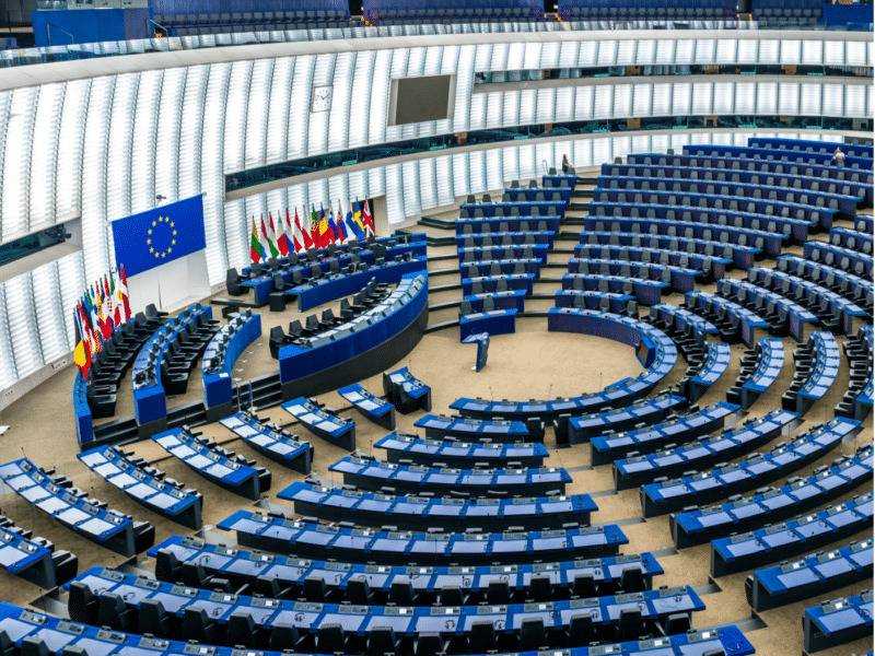 A wide-angle view of the European Parliament's main chamber, featuring its distinctive semi-circular arrangement of seats. The chamber is empty, with rows of blue seats and desks arranged in concentric arcs facing a central podium. A large European Union flag is displayed on the wall behind the podium, alongside an array of flags representing the member states of the EU.