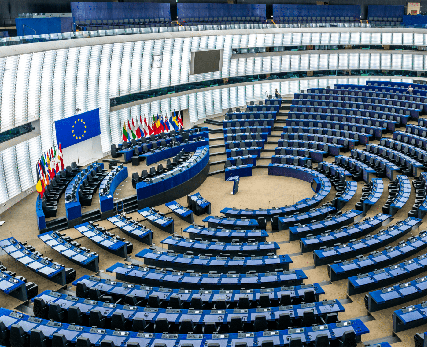 A wide-angle view of the European Parliament's main chamber, featuring its distinctive semi-circular arrangement of seats. The chamber is empty, with rows of blue seats and desks arranged in concentric arcs facing a central podium. A large European Union flag is displayed on the wall behind the podium, alongside an array of flags representing the member states of the EU.