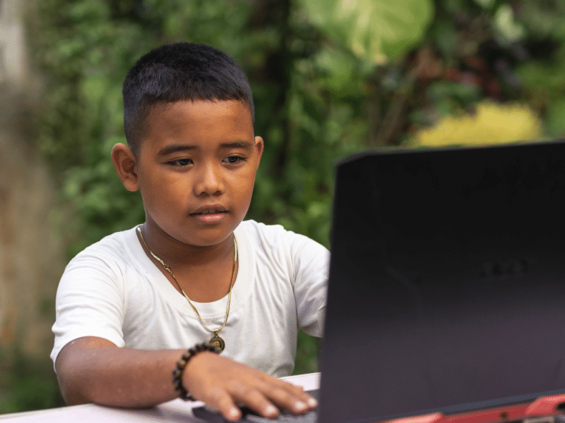 A young Asian boy on a laptop sat on a desk outside (you can see large foliage in the background that suggests this is in Southeast Asia). The camera is positioned so you can see the face of the boy looking at the laptop as he types. He looks inquisitive.