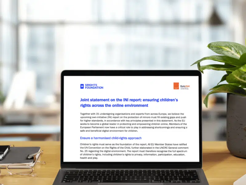 A laptop placed on a wooden desk in a bright workspace displays a joint statement by the 5Rights Foundation and Eurochild regarding the INI report on the protection of minors. The document on the screen features the 5Rights Foundation logo in blue and the Eurochild logo in orange. The title, written in bold blue text, emphasises the importance of protecting minors online. The workspace surrounding the laptop includes a neatly stacked notebook, a pair of glasses, and a green plant beside the laptop, contributing to a professional and organised setting.