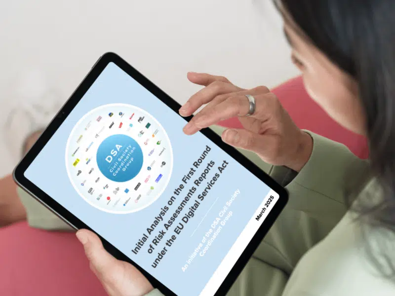 A person holding a tablet displaying a report titled 'Initial Analysis on the First Round of Risk Assessments Reports under the EU Digital Services Act.' The cover features a blue circular graphic in the centre labeled 'DSA Civil Society Coordination Group,' surrounded by logos of the organisations members of the group. The title is written in bold black text on a light blue background, with 'March 2025’ indicated at the bottom.