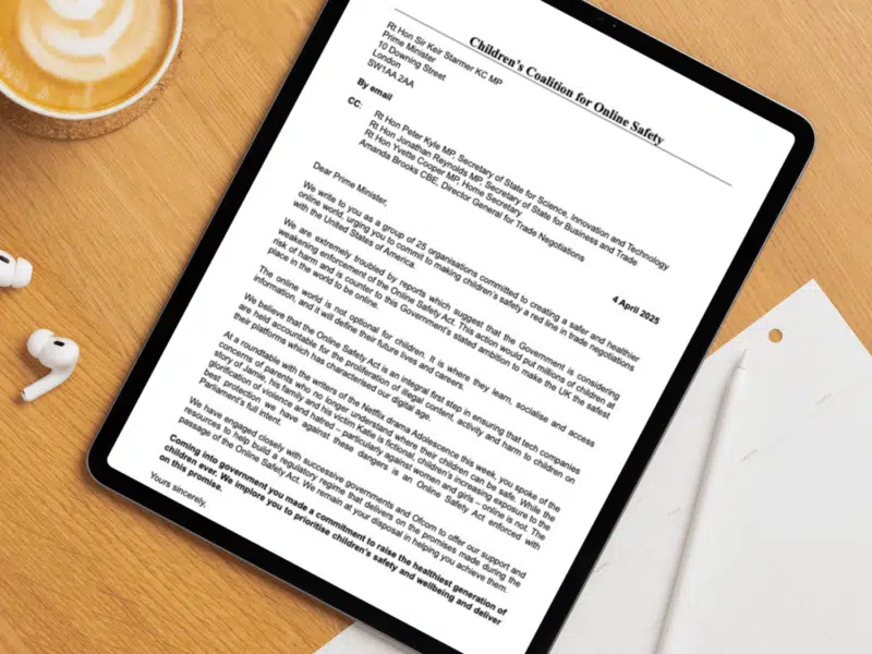 A tablet displaying a formal letter addressed to the UK Prime Minister, titled "Children’s Coalition for Online Safety." The letter, dated 4 April 2025, is shown on a wooden desk alongside a cup of coffee, two white wireless earbuds, and a blank weekly planner with a pen resting on it. The letter emphasises the importance of enforcing the Online Safety Act to protect children online.