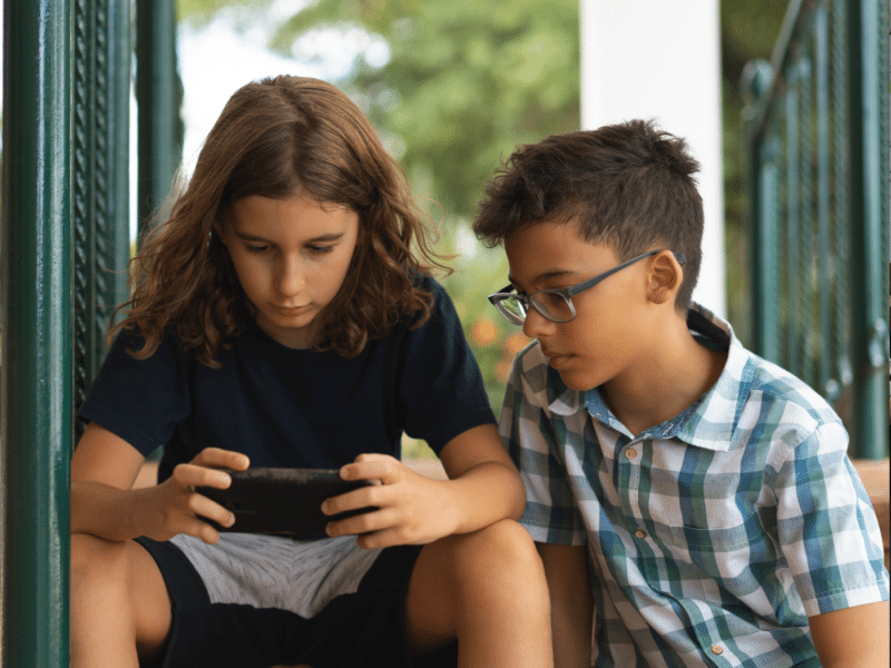 Two children sitting close together on a porch or outdoor steps, with one of them focused on a smartphone. The child on the left, who has long brown hair and is wearing a navy blue t-shirt, is intently looking at the phone he is holding in both hands. The child on the right, who has short dark hair and is wearing glasses and a green and white plaid shirt, is sitting slightly behind and to the side, also looking at the phone. The background shows some greenery, suggesting they are outdoors on a porch or patio. Both children appear to be concentrating on whatever is displayed on the phone, suggesting that they might be playing a game or watching something together.