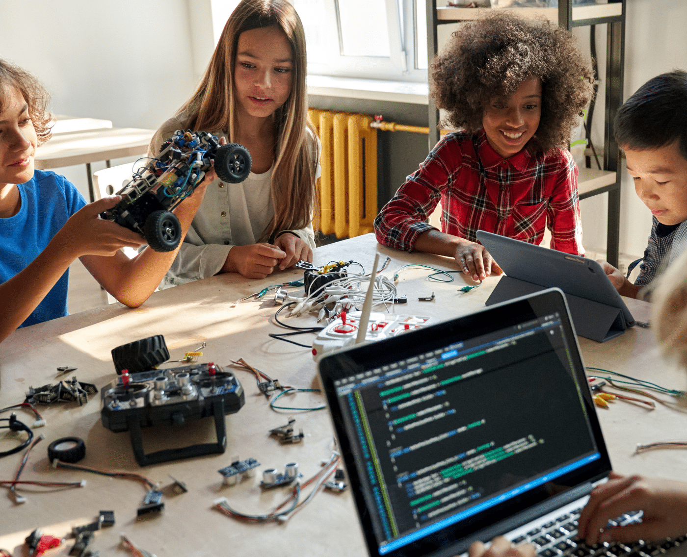 A diverse range of children are sat at a desk, engaging with robotic equipment and technology. They are happy and concentrating.