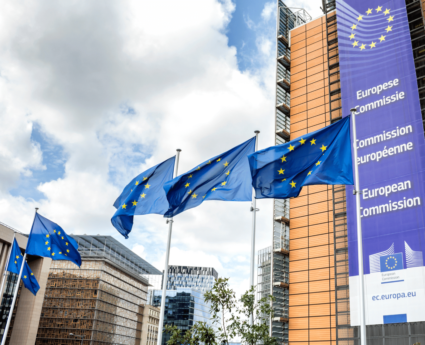 Image of European Commission Headquarters building in Brussels, Belgium, Europe. In the middle of the image are three European flags waving in the wind. To the right is the European Commission building. On the building is a banner which features the logo of the European Commission, and the European Commission translated into multiple languages.
