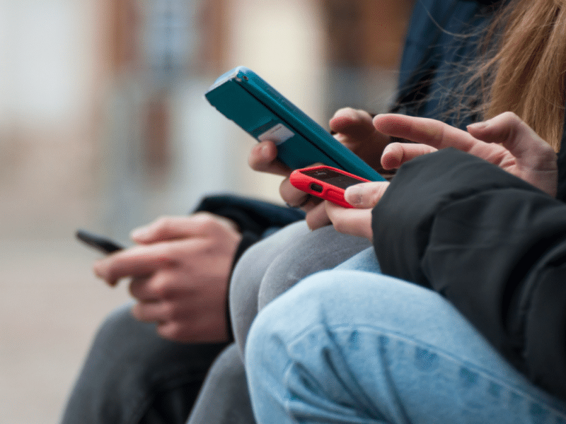 Two white teens are on their phones. The focus of the image is on their hands interacting with the phones, with their faces and upper body not visible to the image.