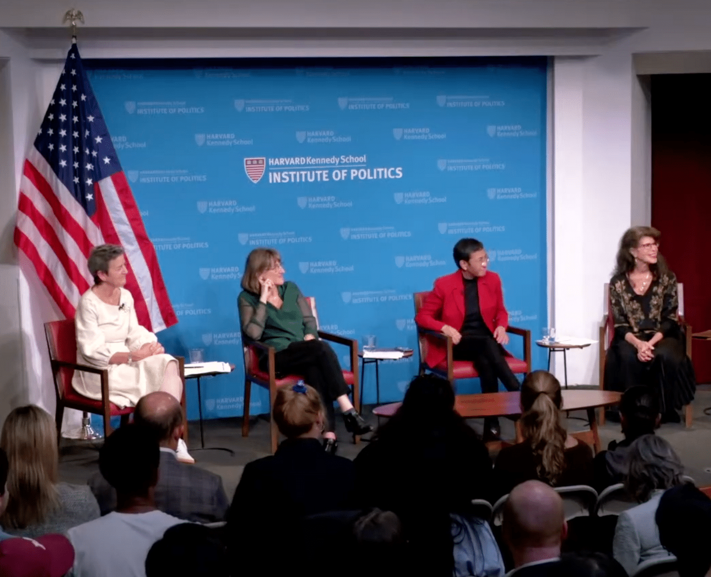 From left: Margrethe Vestager,  Baroness Beeban Kidron,  Maria Ressa, and Shoshana Zuboff. They are sat on a stage. There is a large US flag to the left, behind Margrethe Vestager. The backdrop is a pale blue with the logo and text 'Harvard Kennedy School: Institute of Politics'. 