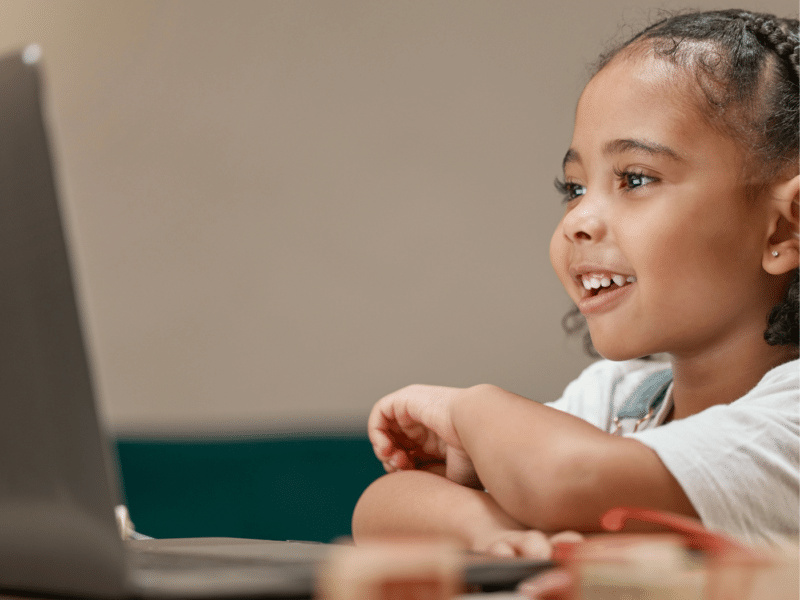 Young black girl on a laptop. Smiling and looking curiously at a laptop. Both the girl and the laptop are side on. The laptop is to the left of the image, and the girl is to the right.