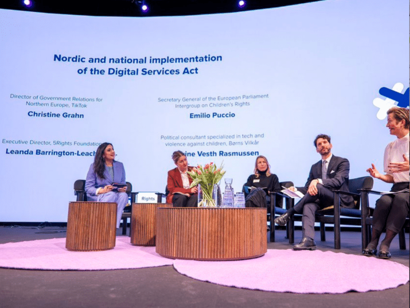 A panel discussion is taking place on a stage with five participants seated in a semicircle. The backdrop displays the title 'Nordic and national implementation of the Digital Services Act' along with the names and titles of the panellists. The panellists are Leanda Barrington-Leach, Executive Director of the 5Rights Foundation; Christine Grahn, Director of Government Relations for Northern Europe at TikTok; Emilio Puccio, Secretary General of the European Parliament Intergroup on Children's Rights and Ane Vester Rasmussen, Political consultant specializing in tech and violence against children at Berns Vilkår. The stage is decorated with a round wooden coffee table in the centre, holding a bouquet of flowers, water bottles, and glasses.