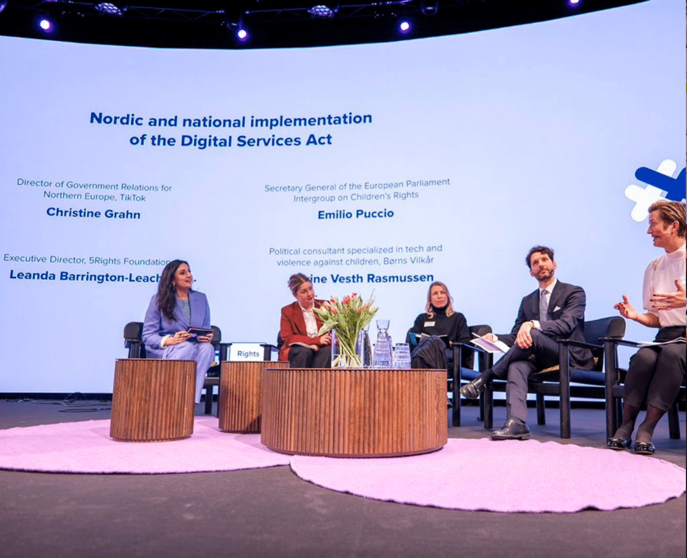 A panel discussion is taking place on a stage with five participants seated in a semicircle. The backdrop displays the title 'Nordic and national implementation of the Digital Services Act' along with the names and titles of the panellists. The panellists are Leanda Barrington-Leach, Executive Director of the 5Rights Foundation; Christine Grahn, Director of Government Relations for Northern Europe at TikTok; Emilio Puccio, Secretary General of the European Parliament Intergroup on Children's Rights and Ane Vester Rasmussen, Political consultant specializing in tech and violence against children at Berns Vilkår.