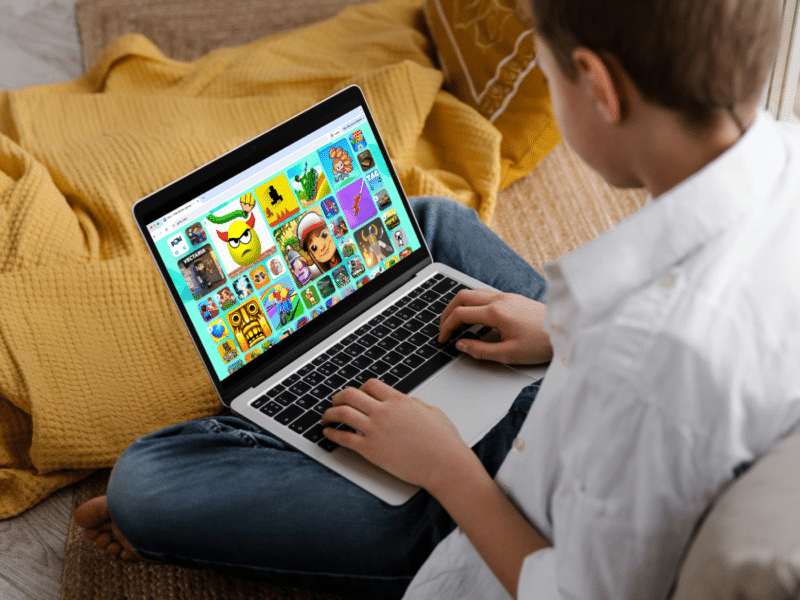 A boy sits cross-legged on the floor using a laptop. The screen displays the homepage of Poki.com, a gaming platform. The page shows a colourful grid of game icons, suggesting he's choosing a game to play. He wears a white shirt and jeans, with a yellow blanket and pillow visible in the background.
