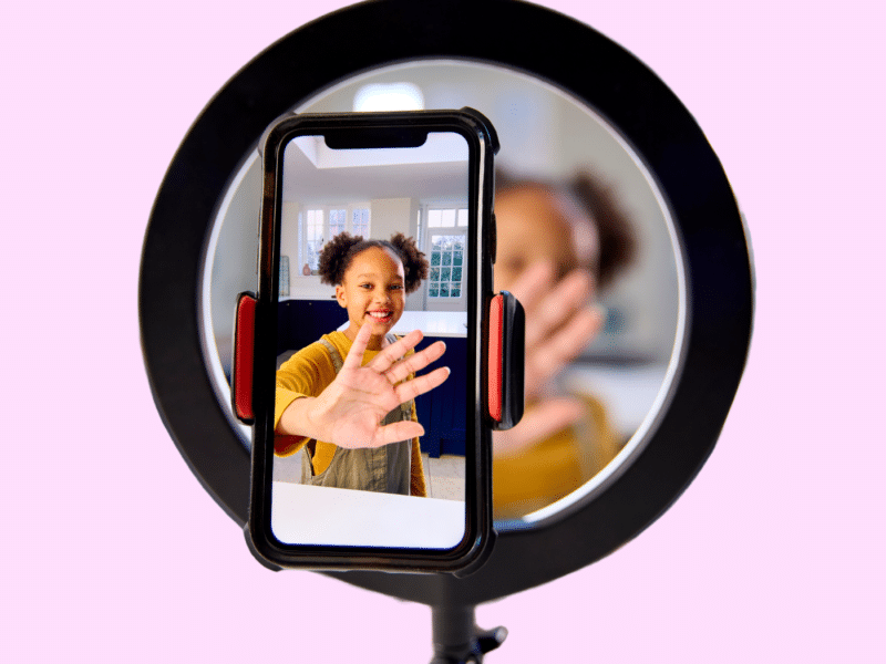 A young girl at home is recording a video with her smartphone to upload on social media. She looks happy and she is wearing brown overalls over a mustard-coloured shirt.