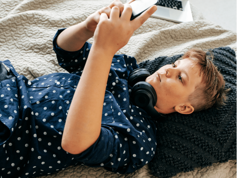 A wider shot of the same scene, featuring a young person lying on a bed. They are wearing a navy blue polka dot shirt, with black headphones resting around their neck. The person is holding a smartphone, gazing intently at the screen while relaxing, with a laptop placed in the background on the bed. This is the full version of the close-up shown above.