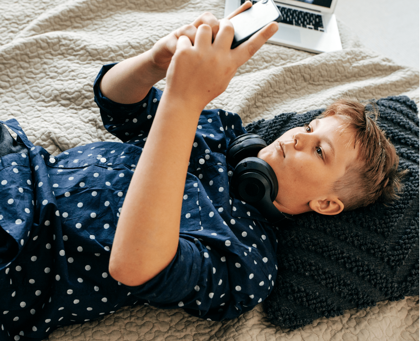 A wider shot of the same scene, featuring a young person lying on a bed. They are wearing a navy blue polka dot shirt, with black headphones resting around their neck. The person is holding a smartphone, gazing intently at the screen while relaxing, with a laptop placed in the background on the bed. This is the full version of the close-up shown above.