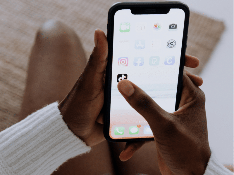 A wider view showing the same scene. A person is sitting, holding a smartphone in both hands, with their thumb about to select the TikTok app. The person is wearing a white long-sleeve top, and the phone screen displays various app icons. This is the full version of the close-up shown above.