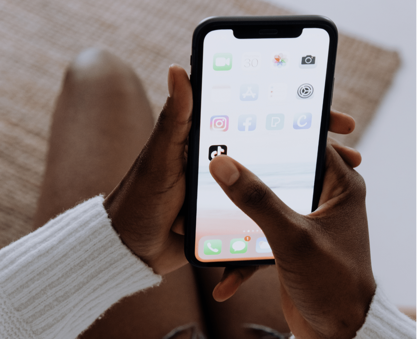 A wider view showing the same scene. A person is sitting, holding a smartphone in both hands, with their thumb about to select the TikTok app. The person is wearing a white long-sleeve top, and the phone screen displays various app icons. This is the full version of the close-up shown above.