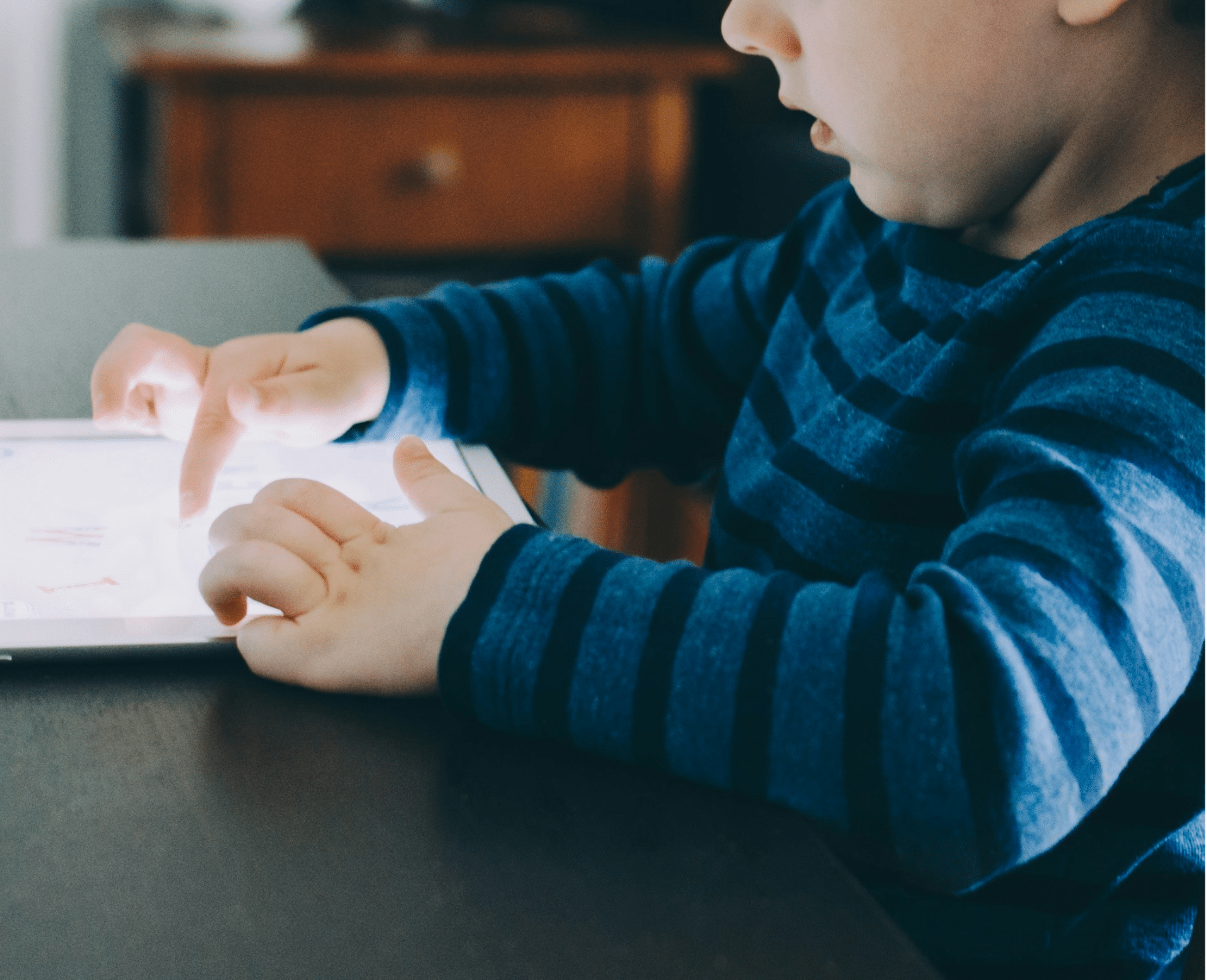 A wider shot showing the same scene in more detail. A young child, dressed in a blue and dark blue striped shirt, is sitting at a table, focused on using a tablet. The child’s fingers are touching the screen, and part of a wooden chair is visible beside them. This is the full version of the close-up shown above.