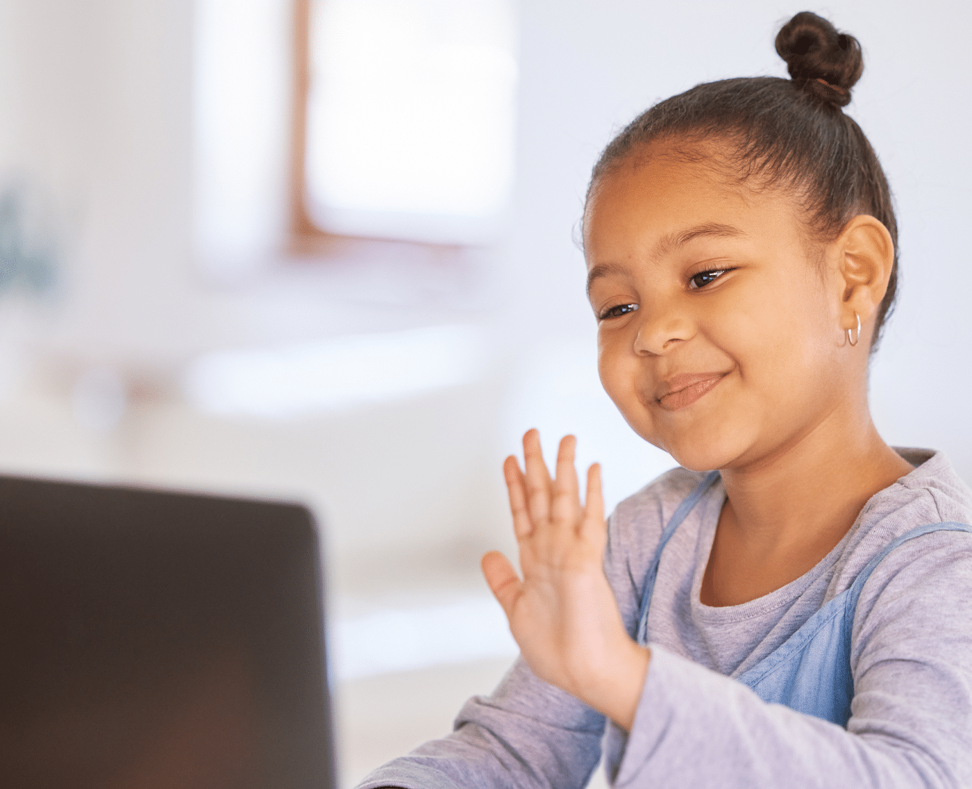 A young black girl on a laptop. She is smiling and waves at the laptop in front of her, which suggests she is on a video call.
